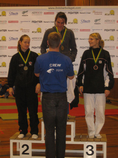 prispall64damvit Mikael Forssén delar ut pris till Ester från Kaisho efter vinst i -64 kg vitbälte. Sara Svensson från Nacka kom tvåa och Siri Fagerlund, Kaisho tog bronset.
